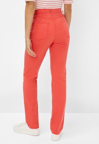 Rote High-Waist-Jeans mit geradem Bein, ausgestattet mit zwei Gesäßtaschen und einem glatten Baumwollmaterial. Getragen mit weißen Sneakers.