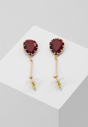 Kultaiset tippakorvakorut, joissa on tippa-muotoiset tummanpunaiset jalokivet ja kirkkaat korvakorujen takakappaleet valkoisella pinnalla.