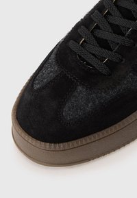 Zapatilla negra que presenta una combinación de materiales de ante y fieltro, puntera redonda, cordones negros y una suela de goma texturizada en marrón.