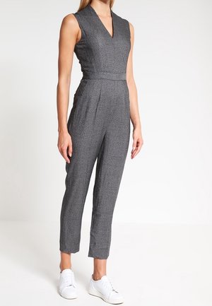 Mujer que lleva un mono sin mangas de color gris oscuro texturizado con escote en V y piernas cónicas, combinado con zapatillas blancas, de pie sobre un fondo blanco.