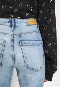 Jean en denim bleu clair avec poches arrière et une étiquette sur la ceinture. Associé à un sweat-shirt noir avec des motifs de cercles dorés.