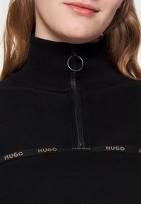 Sudadera negra con cremallera y cuello alto, con la marca HUGO en dorado en el pecho y un tirador de cremallera metálico circular.