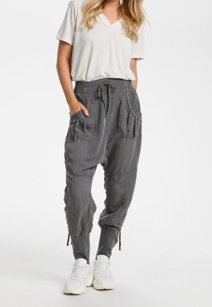 Vrouw die een witte V-hals top met korte mouwen draagt, grijze broek met koordjes en grote zakken aan de voorkant, en witte sneakers. Ze staat met haar handen in haar zakken.