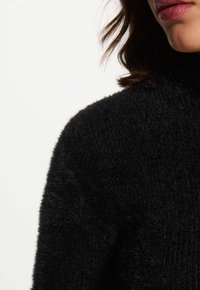 Primo piano di una persona che indossa un maglione nero di pelliccia, con capelli mossi e castani parzialmente visibili su uno sfondo bianco semplice.