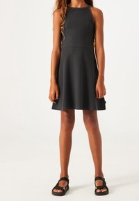 Schwarzes, geripptes Kleid mit eng anliegendem Oberteil und ausgestelltem Rock. Es hat ein ärmelloses Design und eine glatte Textur. Getragen mit schwarzen Sandalen.