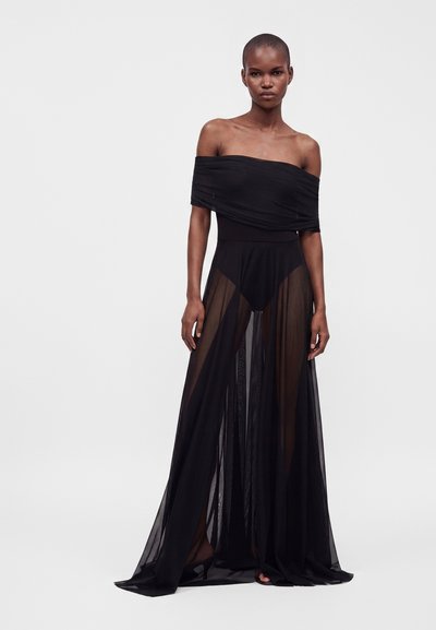 Femme portant un haut noir épaules dénudées avec un pantalon noir transparent taille haute, debout devant un fond uni.