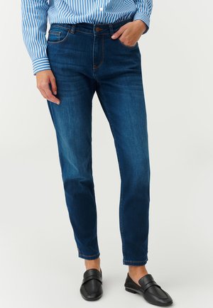 Tmavomodré džíny so slim fit strihom, ktoré majú päť vreciek, zapínanie na gombík a jemné kontrastné švy. Nosia sa s čiernymi poltopánkami.