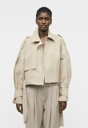 Femme noire portant une veste beige oversize avec de larges poignets et un pantalon plissé taille haute assorti sur un fond uni.