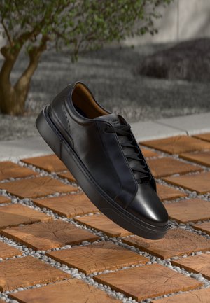 Black leather sneaker with black laces hovering above wooden square tiles separated by small gray stones outdoors.