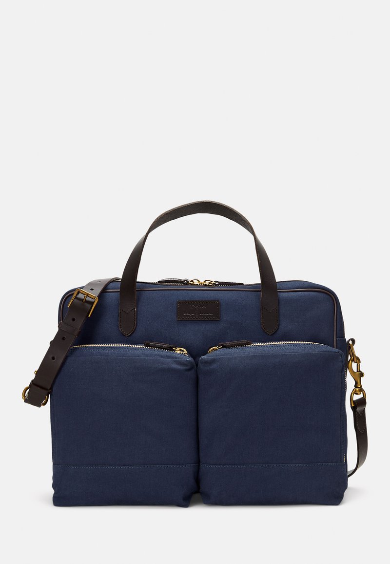 Saco de lona azul-marinho com dois bolsos frontais com zíper, alças de couro castanho e correia ajustável, em forma retangular, textura suave, acabamentos em metal dourado.