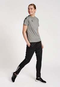 Pelēka sporta T-krekls ar īsām piedurknēm un melnām detaļām, kopā ar melnām konusveida biksēm ar baltām sānu svītrām un gludu tekstūru.