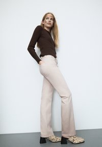 Brązowa, prążkowana bluzka z długim rękawem i detalem guzikowym, zestawiona z jasnoróżowymi dżinsami w formie dzwonów oraz wzorzystymi beżowymi klapkami na blokowym obcasie.
