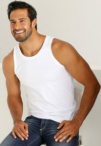 Balta medvilnės marškinėlių su apvaliomis iškirptėmis ir plačiomis petnešėlėmis, derinama su mėlynais džinsais. Modelis sėdi, šypsosi, demonstruodamas marškinėlių prigludimą.