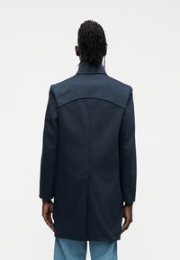 Palton bleumarin cu croială ajustată, guler structurat, detaliu pe yoke-ul din spate și mâneci lungi; asortat cu pantaloni din denim deschis la culoare.