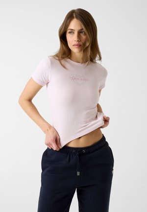 Femme soulevant un t-shirt moulant rose clair avec le texte "GUESS JEANS" sur la poitrine, portant un pantalon bleu marine foncé à cordon, sur un fond uni.