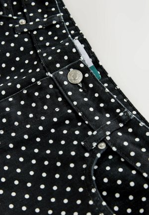 Sorte stofoveraller med hvide prikkede mønstre, udstyret med sølvfarvede metalknapper og kontrasttråd. Nærbillede afslører den strukturerede fløjlsmateriale.