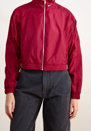 Persona con chaqueta Nike roja con cremallera y logo blanco y pantalones negros, de pie con las manos relajadas a los lados.