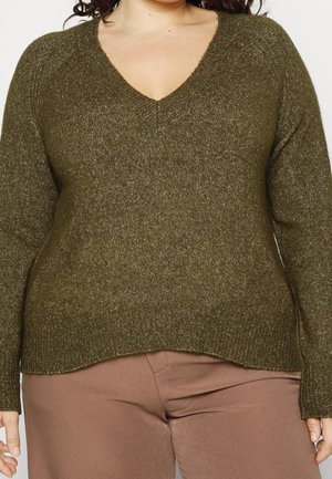 Žena v olivově zeleném pleteném svetru s výstřihem do V a světle hnědých kalhotách, stojící před jednobarevným pozadím.