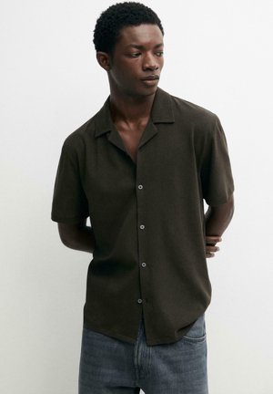 Uomo con camicia marrone scuro a maniche corte e jeans blu, guarda a sinistra con le mani dietro la schiena su sfondo bianco.