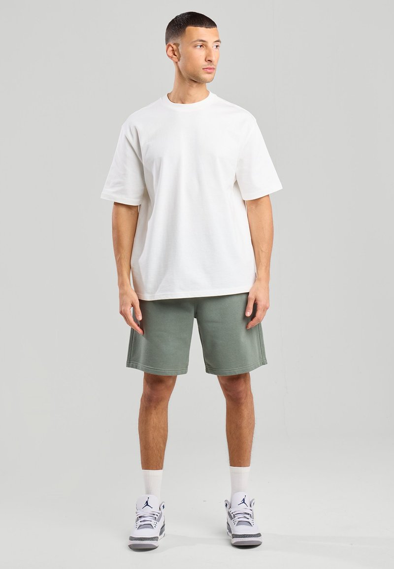 White cotton t-shirt with short sleeves paired with green cotton shorts. Model wears white sneakers and white crew socks. Plain design.
