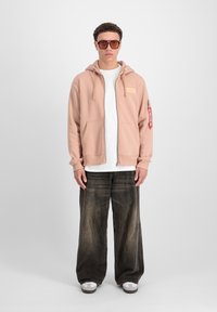 Felpa con zip di colore rosa chiaro, indossata su una maglietta bianca, abbinata a pantaloni larghi in jeans sbiaditi e scarpe slip-on grigie. Gli occhiali da sole marroni completano il look.