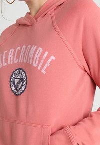 Růžová mikina z měkké tkaniny, vybavená klokaní kapsou, kapucí se šňůrkou a potištěným textovým logem vpředu.