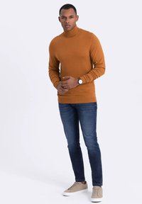 Brauner Rollkragenpullover, schmal geschnittene blaue Jeans und hellbraune Sneakers. Der Pullover hat gerippte Bündchen und eine glatte Textur.