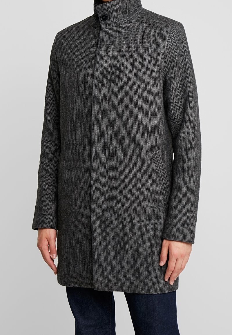 Homme portant un manteau en laine gris foncé à motif chevrons avec un col montant et une fermeture à boutons, associé à un jean bleu foncé.
