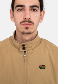Tan zip-up jacket with a high collar. Features black buttons and a small embroidered patch with colorful flowers on the chest.