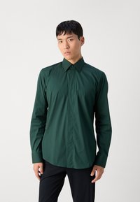 Camisa de manga larga verde oscuro con cuello puntiagudo, con una textura suave y un diseño ajustado. Pantalones negros visibles.