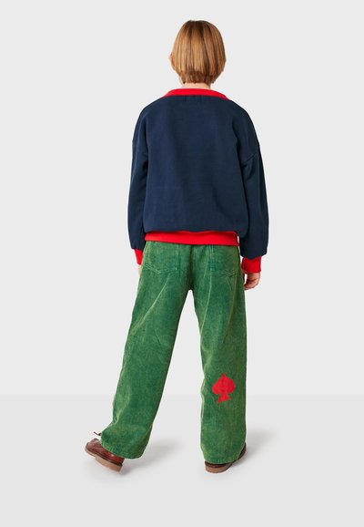 Suéter oversize azul marino con puños rojos, combinado con pantalones de pana verde que tienen un logo de trébol rojo, visto desde detrás.