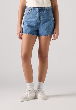 Person iført midtblå denimbukser, hvide sokker og hvide sneakers, der står mod en ensfarvet lys baggrund.