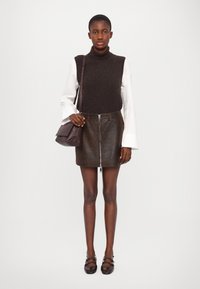 Maglione a collo alto marrone sopra una camicia bianca, mini gonna in pelle marrone con zip frontale, e borsa scura. Modella che indossa scarpe nere con cinturini.