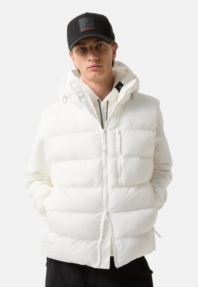 Witte gewatteerde vest met capuchon, voorzien van een ritsluiting, horizontale stiknaden en zijzakken met rits. Gecombineerd met een zwarte pet.