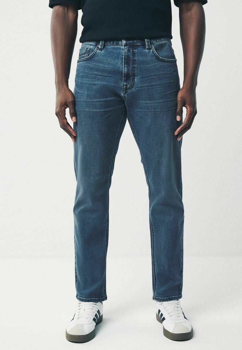 Jean en denim bleu foncé avec une coupe décontractée, présentant un léger délavage, un design à cinq poches et des accents cousus, associés à des baskets blanches et grises.