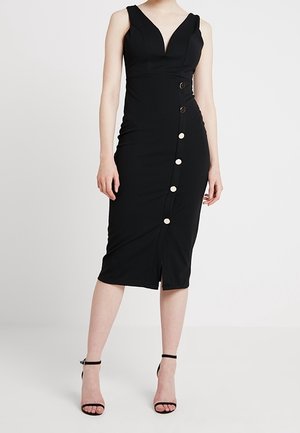Femme portant une robe midi noire sans manches avec un col en V et un détail asymétrique de boutons sur le devant, associée à des talons noirs à brides à la cheville.