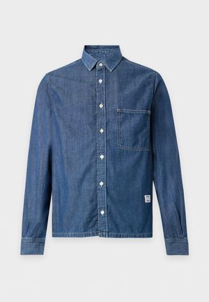 Un hombre luce una camisa vaquera azul con botones, unos pantalones cargo marrones holgados y zapatos negros sobre un fondo blanco liso.