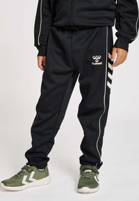 Pantalones de chándal negros con ribete blanco y marcas. Tejido duradero, cinturilla elástica y bolsillos laterales. Combinados con zapatillas verdes que tienen un acento blanco.