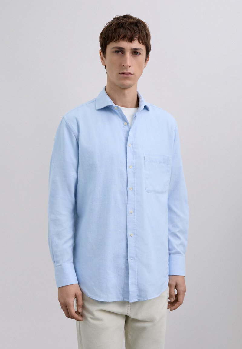 Chemise bleu clair à manches longues en coton, avec un col boutonné, une poche devant et des boutons jaunes. Texture lisse.