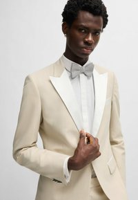 Giacca smoking beige con rever bianchi a punta, abbinata a una camicia bianca e un papillon argento. Tessuto morbido, silhouette aderente.
