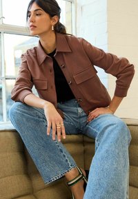 Braune, kürzere Jacke mit großen Vorderaschen, getragen über einem schwarzen Oberteil. Kombiniert mit hochgeschnittenen, hellblauen weiten Jeans und schwarzen Sandalen mit Absatz.