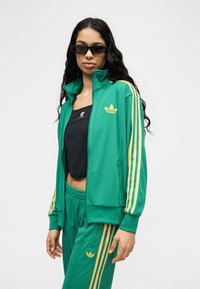 Γυναίκα που φοράει πράσινη φόρμα Adidas με κίτρινες ρίγες και μαύρο crop top, ποζάροντας με γυαλιά ηλίου μπροστά σε απλό φόντο.