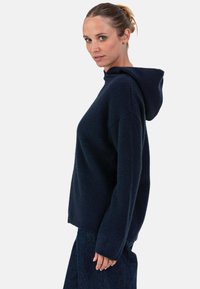 Sweat à capuche bleu marine avec une texture douce, des manches larges et une coupe décontractée. Comprend une capuche à cordon de serrage et un ourlet côtelé, associé à un denim foncé.