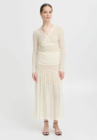 Abito in pizzo avorio con maniche lunghe, design avvolgente e lunghezza maxi fluida. Presenta un motivo testurizzato e un leggero volants all'orlo.