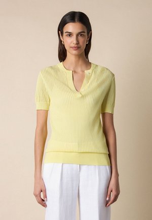 Donna che indossa una maglietta a maniche corte di maglia giallo chiaro con bottoni e pantaloni bianchi, in piedi contro uno sfondo beige semplice.