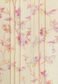 Vyšívaná látka v měkké béžové barvě s květinovými vzory v růžových, žlutých a stříbrných nitích; má vertikální záhyby a hladkou texturu.