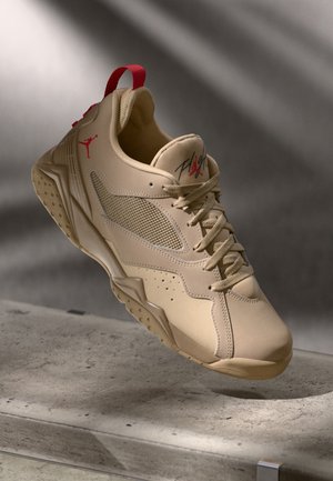 Baskets athlétiques beiges avec logo Jumpman rouge et texte "Flight" sur la languette, coutures détaillées, panneau latéral en mesh et languette rouge au talon.