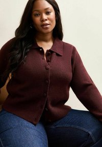 Bordowy kardigan z fakturowanej tkaniny, z guzikami z przodu i klasycznym kołnierzem. Noszony na ciemnoniebieskich dżinsach. Prosty i dopasowany krój.