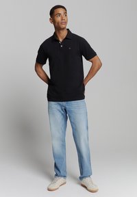 Zwart polo t-shirt met korte mouwen, met een kraag en een klein logo. Gecombineerd met lichtblauwe jeans en beige sneakers.