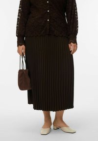 Blouse en dentelle marron foncé à manches longues avec ourlet festonné, jupe midi plissée marron foncé, mules crème et petit sac à main en suède marron.